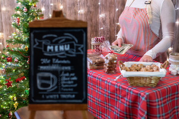 Christmas pastry shop. Woman seller, waitress counting money selling gingerbread, marshmallows, cookies, sweets in a small cozy cafe. Homemade bakery menu in the foreground