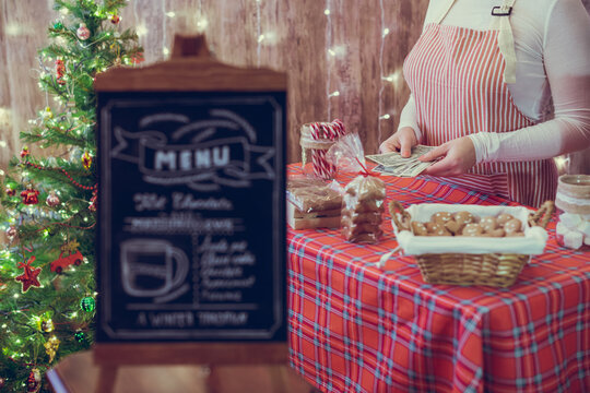 Christmas pastry shop. Woman seller, waitress counting money selling gingerbread, marshmallows, cookies, sweets in a small cozy cafe. Homemade bakery menu in the foreground