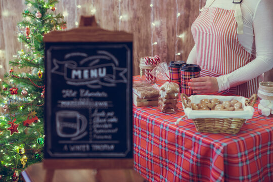 Christmas pastry shop . Woman seller, waitress selling gingerbread, marshmallows, cookies, sweets and hot chocolate in a small cozy cafe. Homemade bakery menu in the foreground