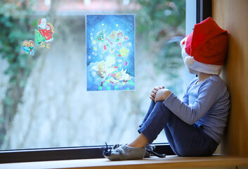 Ein kleiner Junge sitzt mit Weihnachtsmütze am Fenster