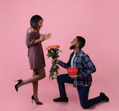 Full Length Portrait Of Romantic Black Guy Giving His Girlfriend Flowers And Gift For Valentine's Day On Pink Background