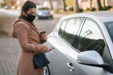 Woman in black face mask hold cuf of coffee by the car and look car key. Woman open car. Protectiv mask in quarantine. Cov-19