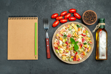 top view of bowl of veggie salad with fork oil bottle tomatoes pepper and notepad on side on dark grey background