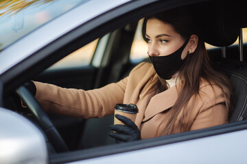 Woman with face mask driving her car with cup of coffee during coronavirus pandemic, Covid-19