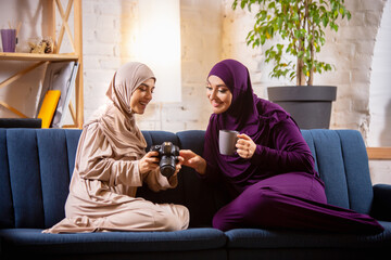 Tea time. Happy and young two muslim women at home talking, smiling, having fun, sitting on sofa. Friendship, modern tech, emotions concept. Lifestyle and culture, arabian traditions, ethnicity.