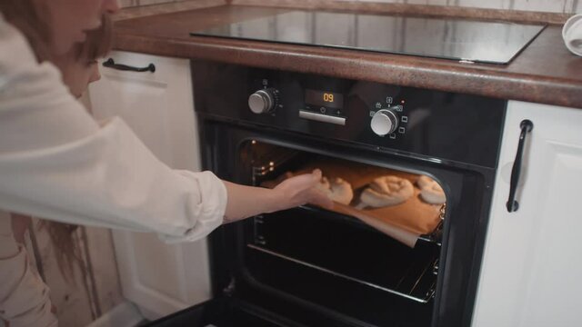 Medium close-up of young mother and daughter in cooking aprons placing baking sheet with homemade pretzels in preheated oven ready for baking
