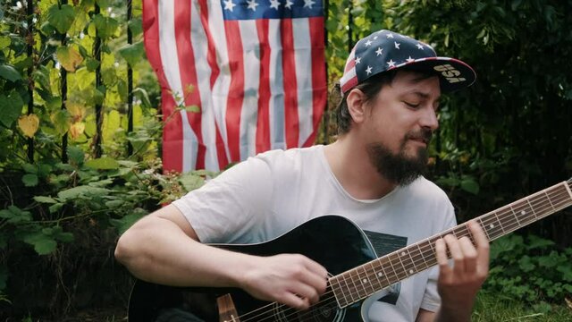 Man in a USA hat playing the guitar with an American flag in the background