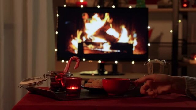 winter holidays and hygge concept - hand placing cup of coffee or hot chocolate to table with ginger cookies, candle, candy canes and gift boxes over tv monitor used as fireplace at cozy home