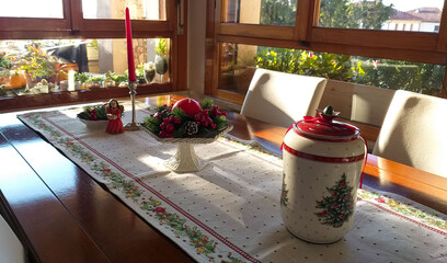 Table with Christmas decorations. Embroidered centerpiece, a painted white ceramic cookie jar, a candle holder, a red candle, an angel. Soft and delicate light that filters through the windows.