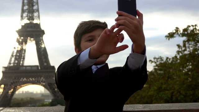 A cheerful teenage boy in a black suit is taking a photo on the phone on the background of the Eiffel tower at the sunrise, Paris, France