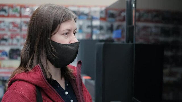 Caucasian a woman wearing a mask against the virus buys a tv in a home appliance store. The woman looks at the LED panel, smiles and says something