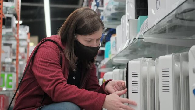 Caucasian a women wearing antivirus medical mask and red jacket picks out portable heater in electronics shop. Going to showroom during pandemic