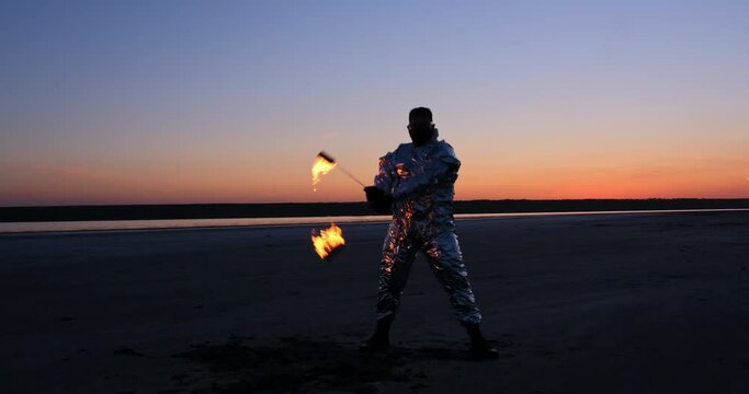 A fiery spinner in a silver suit spins torches on chains burning in flames against the backdrop of the setting sky.