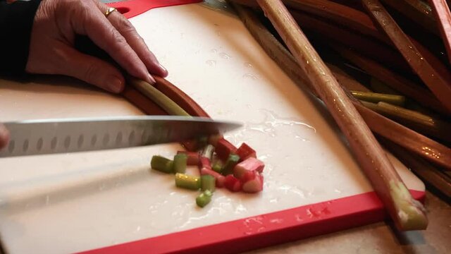 Hands of chef using a knife to cut rhubarb on a red and white cutting board. Slow motion clip with subtle zoom in.