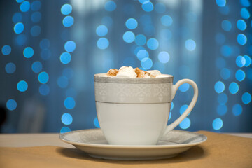 White with silver cup of hot and aromatic cocoa with marshmallow on blue bokeh fairy lights background. Enjoying hot coffee in cozy cafe atmosphere, barista treat or tea time concept.