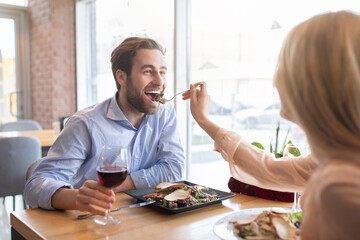 Positive young couple having romantic festive dinner at cafe, celebrating birthday, feeding each other, drinking wine