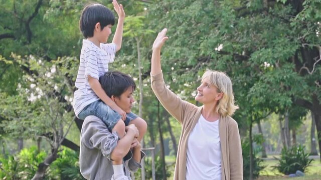 Asian boy sitting on father's shoulder high five with foreigner senior woman in the park. kid learning to greeting with new friend. diversity people friendship concept