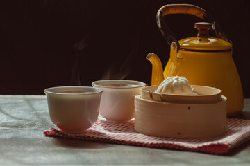 Steamed buns in wooden basket and two cups and yellow pot has a fragrant tea with steam, Morning tea on black background,Food that provides warmth to body and has health benefits in the winter season.