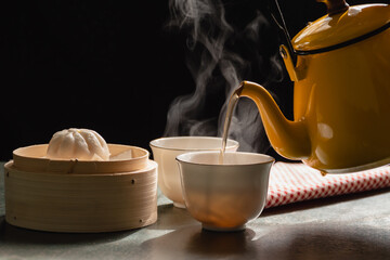 Steam from two cups and yellow pot has a fragrant tea and steamed buns in wooden basket, Morning tea on black background,Food that provides warmth to body and has health benefits in the winter season.