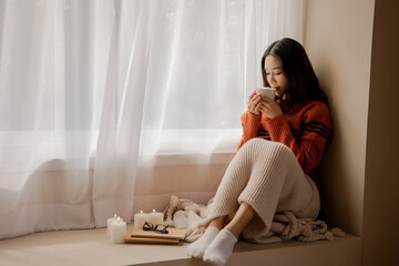 Young beautiful Asian woman holding cup of coffee and marshmallows on top, sitting at home and looking out the window. Happy girl drinking chocolate in sweater in cold weather winter - 401007969