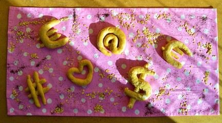 View of a table with different shaped breads: euro, dollar, heart, at, hashtag on a pink tablecloth with white polka dots that symbolizes the relationship between food, the economy and technology