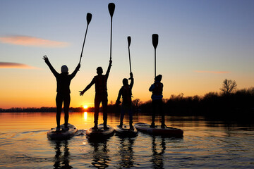 Four friends on stand up paddle board (SUP) on a flat quiet winter river at sunset raising his...