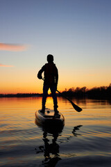 Rear view on silhouette of boy on stand up paddle boarde paddling at sunset on river at cold time