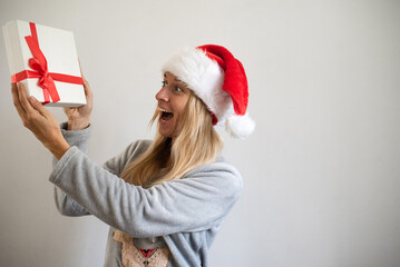 Woman on grey background wearing Christmas pyjama and cap holding a gift. Happy to get and give the gift. Feel fairy tale and magic. new year emotional and mood. ready to Christmas