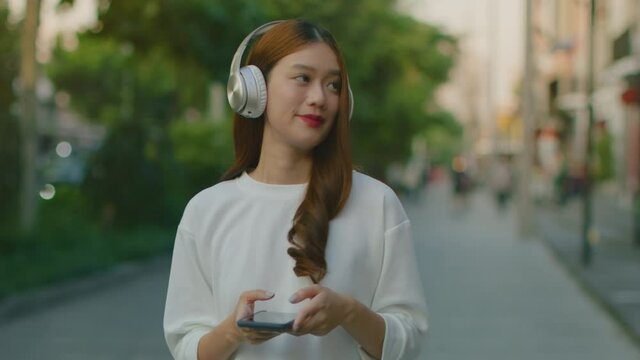 Slowmotion of Happy Young Asian Woman in headphones walking in the city street outdoor having fun listening to music. Joyful Asian Girl in casual walking in the street outdoor. 