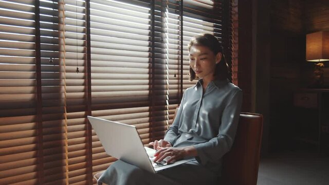 Zoom out shot of young attractive Asian businesswoman sitting near window with blinds in home office and working on laptop