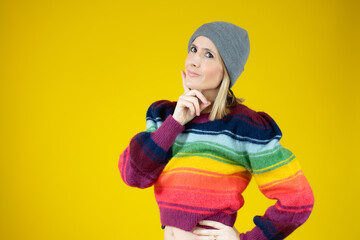 Young woman in winter clothes with doubtful gesture standing over yellow background.