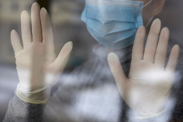 A woman in a medical mask and rubber gloves near the window, self-isolation during the coronavirus.