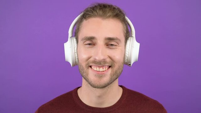 portrait of attractive laughing long-haired man wearing large white wireless headphones. on an isolated purple background. the man is wearing a burgundy sweater. 4K