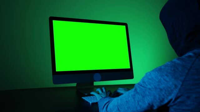 Back view of hacker programmer wearing a hood, typing on keyboard and a green screen display. Unrecognizable man hacking in a dark room. Concept of dark internet, chroma key mock up.