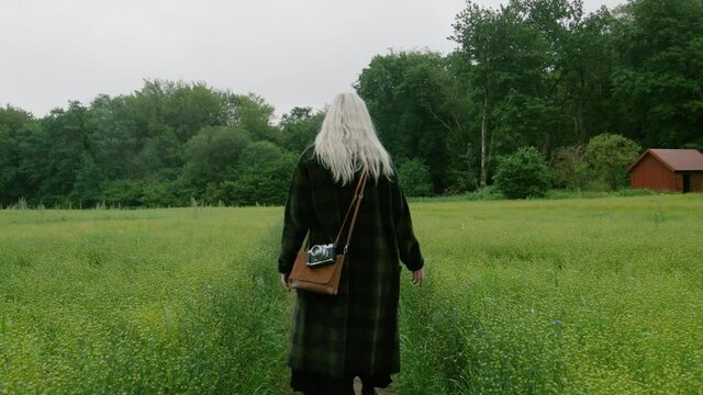 Blonde cute German girl walking across a field of flowers in a black coat vintage bag and camera towards a forest on misty cloudy Scandinavian spring touching with hands wanderlust exploring adventure