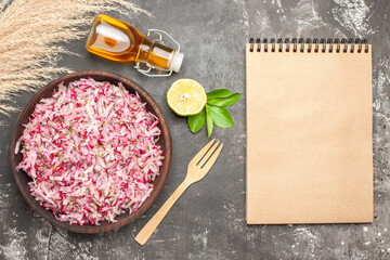Above view of vegetable salad with various ingredients in a brown bowl lemon oil bottle fork and notebook on dark background