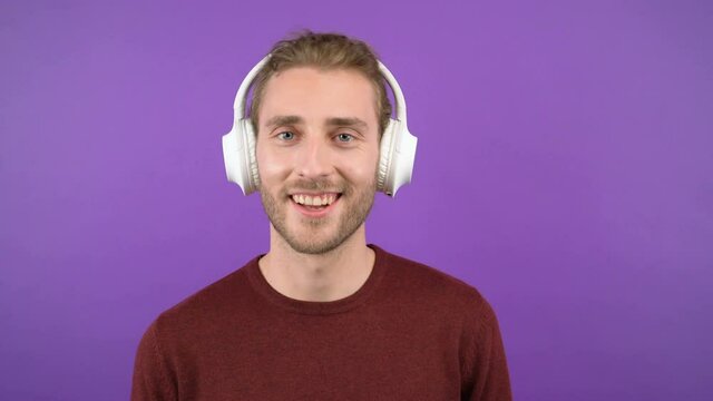 close-up portrait of long-haired attractive man in big white headphones dancing man on isolated purple screen. The man is wearing a burgundy warm sweater. 4K