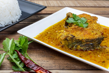 Bengali cuisine - doi katla (catla fish curry with yogurt) in a plate served with white rice with dry red chilli and coriander leaves on wood background.