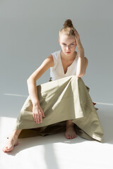 The model girl sits in a long green skirt and white tank top and looks straight. Her elbow is on...