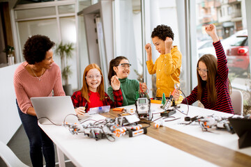 Happy kids with their African American female science teacher with laptop programming electric toys and robots at robotics classroom