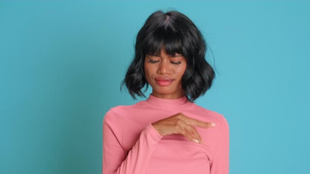 Young dissatisfied dark skinned female model wears casual pink jumper waves hand near nose feels unpleasant smell squints because of stench doesnt like something disgusting over blue studio background