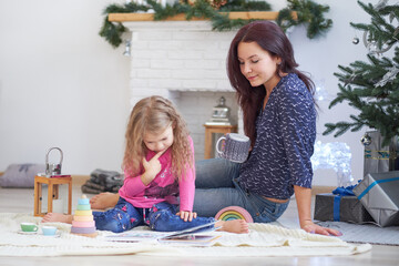 Positive cute young woman mom sits on floor in a festive studio and plays with little girl.. Good Christmas mood concept