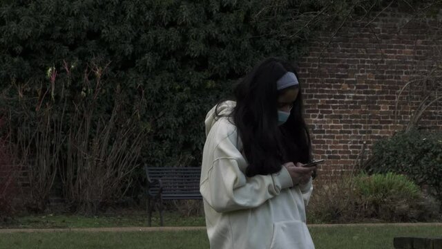 Female Teeanger Wearing Face Mask Checking Phone In Public Park During Lockdown. Locked off