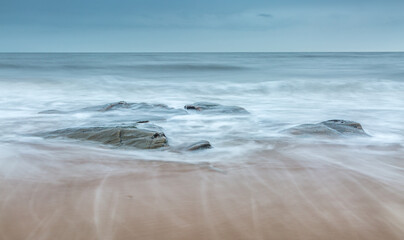 A group of rocks on the beach as the wave backwash returns to the sea over the sand on a cloudy day (1)