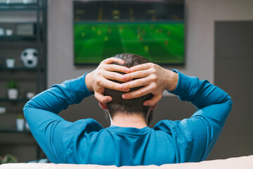 Back view of yong bearded man watching tv and holding his head with hands stressed about football...