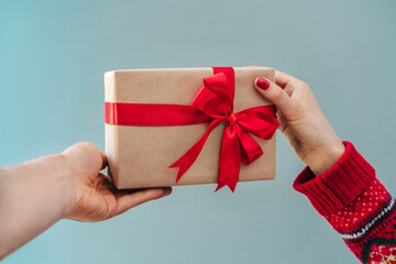 Close up cropped shot of two male and female hands sharing present wrapped in craft paper and decorated with red satin ribbon with each other