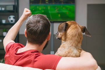 Back view portrait of a man sitting at home on sofa with his dog friend watching football play live broadcast on tv together