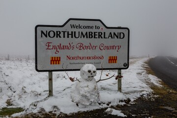 A frightening looking snowman next to the Northumberland border sign, with snow on the ground
