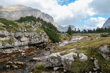 Fototapeta na wymiar A rushing torrent in Italian Dolomites. There is not much water in the torrent, a few stones are popping out. High mountain chains in the back. Forest on the side. Power of the nature