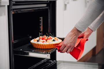 Women's hands reach for a beautiful, freshly made berry pie from the oven in a white modern kitchen.